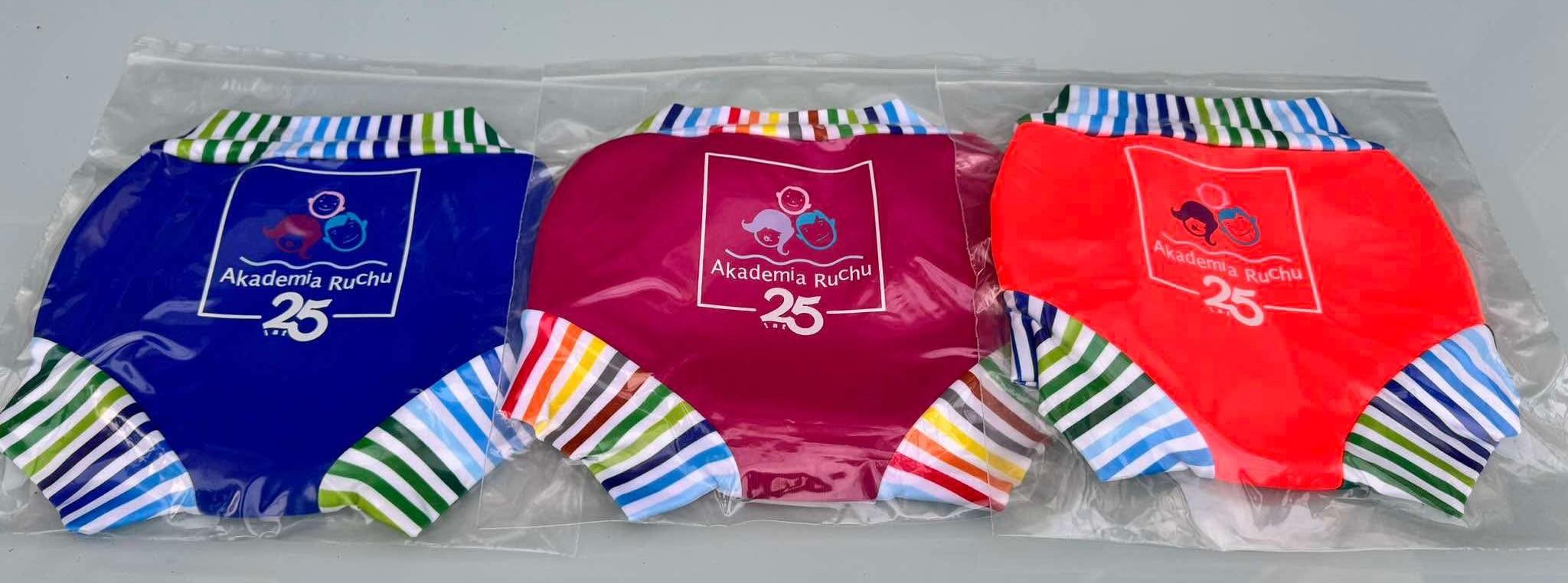 Trzy pary majtek neoprenowych Akademii Ruchu w opakowaniach — granatowe, różowe i czerwone, z kolorowymi paskami przy nogawkach i nadrukiem logo z okazji 25-lecia